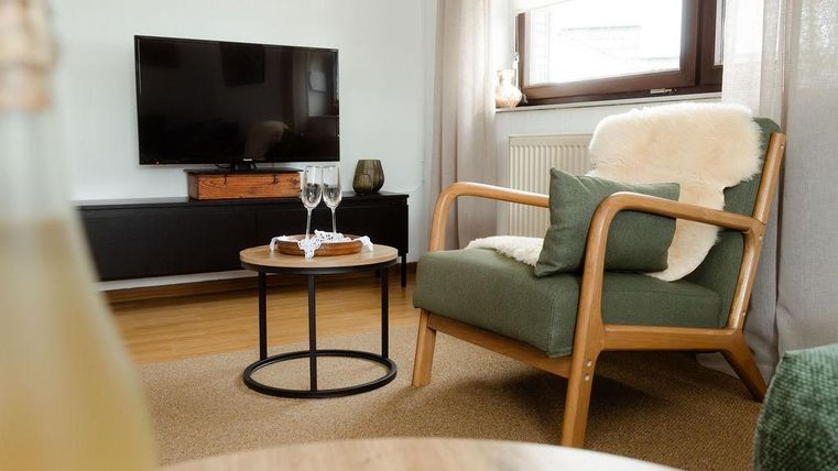 Un espace de vie confortable avec un fauteuil douillet et une table basse. À l'arrière-plan, on peut voir une télévision et des fenêtres laissant entrer la lumière du jour.