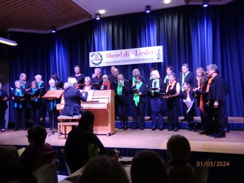 A choir performs on a stage while a pianist accompanies. The singers wear various colored scarves and present their performance to an audience.