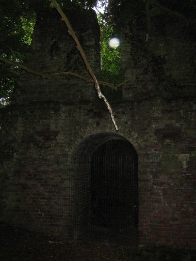 An old, partially weathered ruin made of brick, surrounded by trees and dense vegetation. The entrance of the ruin is closed off with a grille and the atmosphere feels mysterious and gloomy.