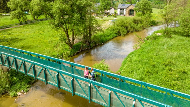 Two people on a green bridge over a river in a rural landscape.