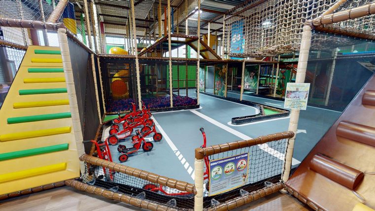 Een kleurrijk ingericht indoor-speelparadijs met verschillende speelgebieden. Er zijn een glijbaan, klimelementen en een strafruimte voor kinderen met voertuigen.