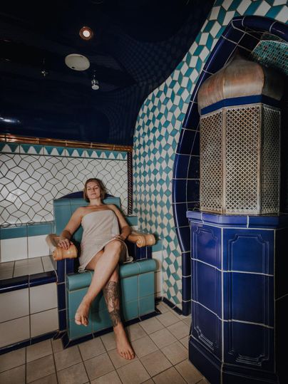 Een persoon zit in een blauwe wellnessruimte op een comfortabele stoel. De muren zijn versierd met kleurrijke tegels die een ontspannen sfeer creëren.