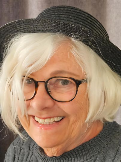 Eine lächelnde Frau mit grauem Haar trägt eine schwarze Mütze. Sie trägt eine graue Strickjacke und schaut freundlich in die Kamera.