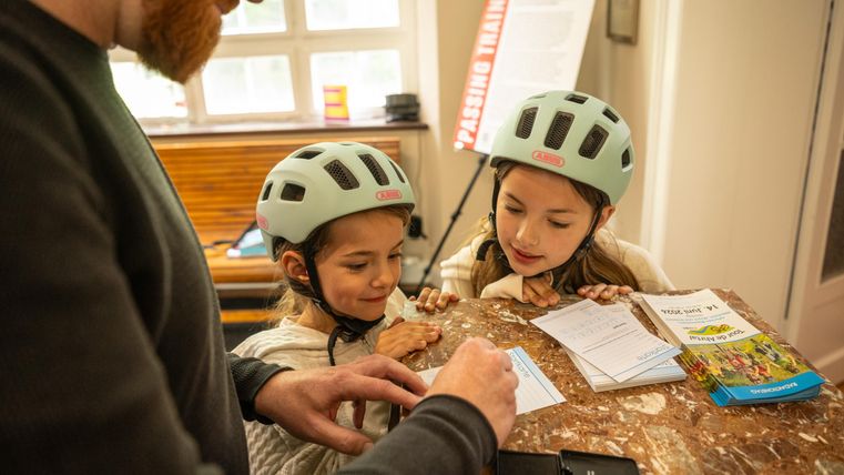 Twee kinderen met fietshelmen kijken nieuwsgierig naar een tafel, terwijl een volwassene iets op papier schrijft. De sfeer is vriendelijk en uitnodigend.