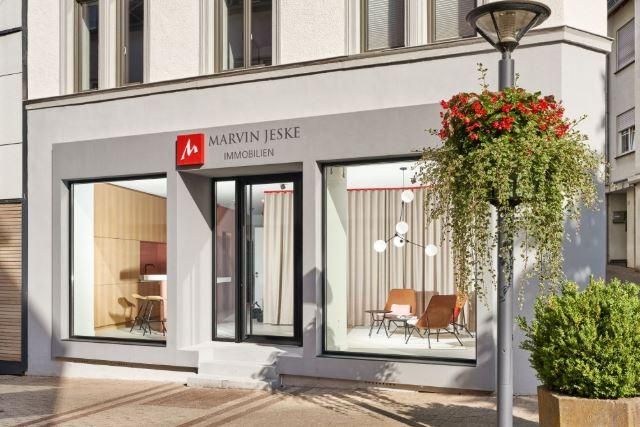 Ein modernes Immobilienbüro mit großen Fenstern und klaren Linien. Die Fassade ist hell und einladend, umgeben von grünen Pflanzen.