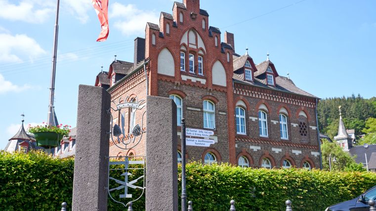 Un bâtiment historique avec une façade rouge frappante et de belles fenêtres. Au premier plan, une clôture et un chemin sont visibles, entourés de buissons verts.