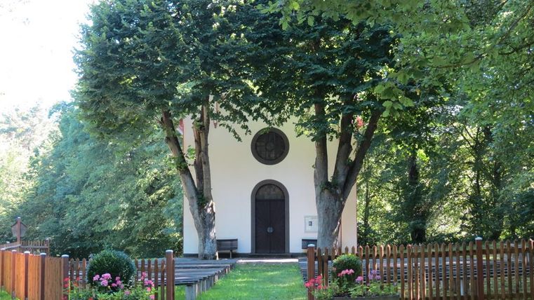 Une petite chapelle entourée de grands arbres et de fleurs colorées. Le chemin menant à la chapelle est bordé d'une clôture en bois.
