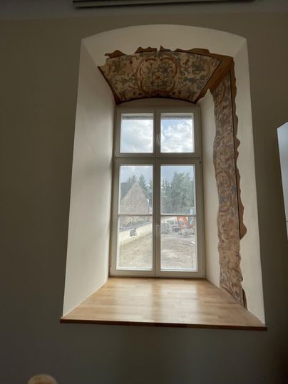 Une fenêtre avec une planche en bois et un cadre décoratif. À l'arrière-plan, on peut voir des arbres et des chantiers.