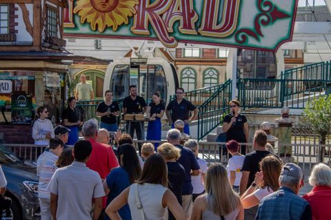 Un groupe de personnes se tient devant une attraction colorée tout en faisant une présentation. De nombreux spectateurs écoutent avec intérêt et le ciel est dégagé.