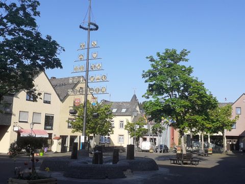 A quiet place with a maypole and several trees. In the background, pretty buildings and a clear blue sky can be seen.