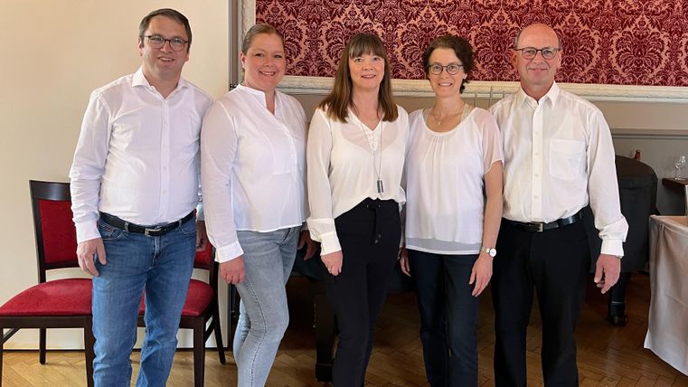 Eine Gruppe von fünf Personen steht in einem eleganten Raum mit rotem Tapetenmuster. Alle tragen weiße Oberteile und lächeln freundlich in die Kamera.