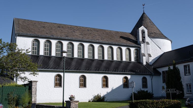 Église blanche avec tour ronde et toit pointu, entourée d'espaces verts, sous un ciel bleu.
