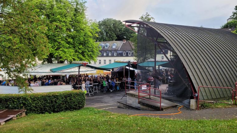 Eine Veranstaltung im Freien mit vielen Gästen unter einem Zeltdach. Im Hintergrund sind grüne Bäume und ein Gebäude sichtbar.