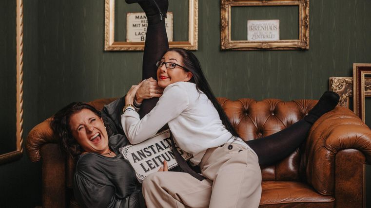 Two women are lying on a brown leather sofa. One of the women is holding a sign that says "Institution management" and is laughing, while the other is lying with one foot up on the sofa.