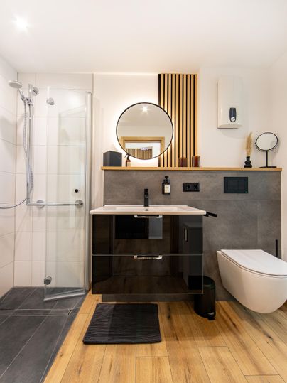 A modern bathroom with a shower, a washbasin, and a flushable toilet. The floor is wooden, and there is a large mirror as well as design elements made of wood and marble.