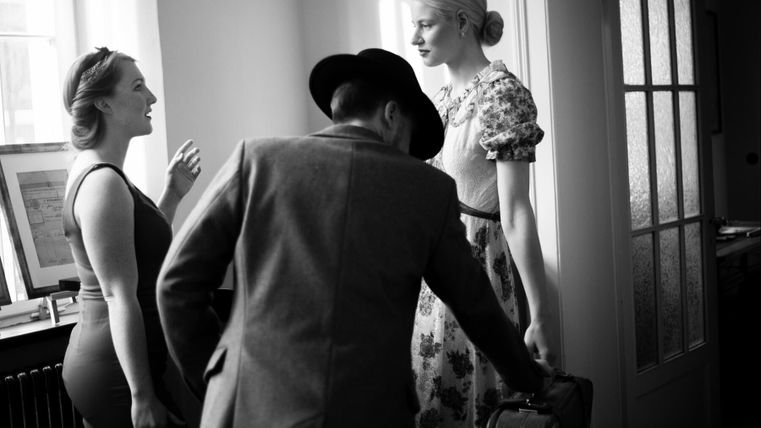 Trois personnes dans une pièce lumineuse, deux femmes discutent tandis qu'un homme les écoute. L'une des femmes porte un motif floral, l'autre une tenue élégante.