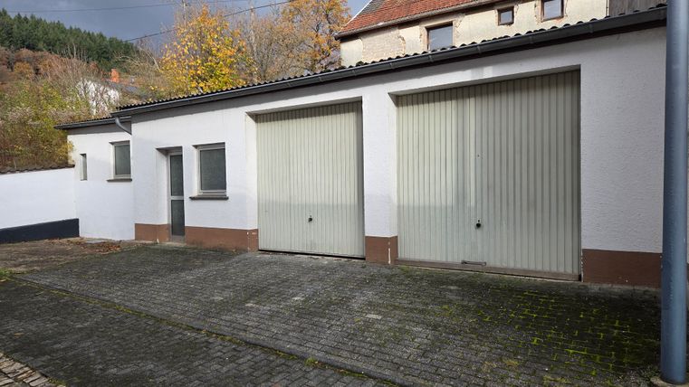 Un bâtiment de garage avec deux grandes portes de garage grises. À l'arrière-plan, des arbres et un autre bâtiment sont visibles.