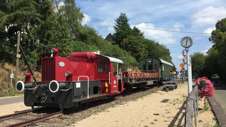 Ancienne gare de Pronsfeld avec une locomotive rouge et de vieux wagons sur des rails, entourés d'arbres et de fleurs.