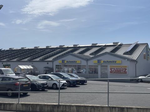 Un bâtiment commercial nommé "Schmitz" avec un toit moderne. Devant le bâtiment, plusieurs voitures sont garées sur un grand parking.
