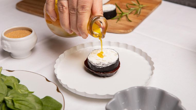 Een hand giet honing over een klein, gelaagd gerecht op een wit bord. Op de achtergrond zijn verse ingrediënten en een houten snijplank zichtbaar.