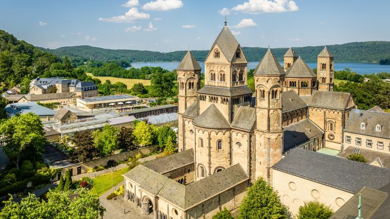 Een indrukwekkende abdij met een gotische architectuur, omgeven door groene bomen. Op de achtergrond is een rustige meer en een blauwe lucht te zien.
