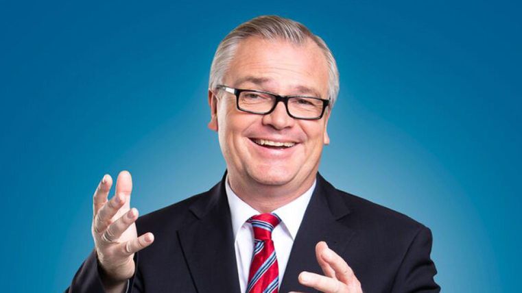 Ein lächelnder Mann in einem Anzug mit einer Krawatte. Der Hintergrund ist blau und er gestikuliert freundlich.