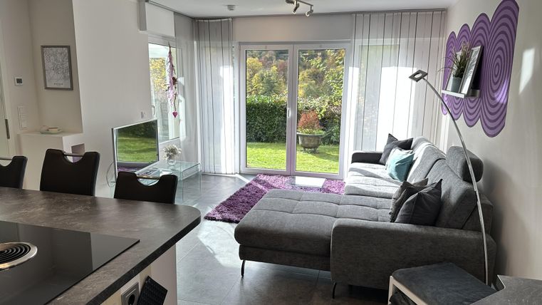 A modern living room with a gray sofa and large windows that provide a view of the garden. The space is bright and friendly designed, with a stylish rug and decorative elements.