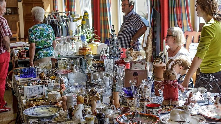 Ein lebhafter Flohmarkt mit vielen verschiedenen Sammlerstücken und Geschirr. Menschen stöbern durch die Stände und unterhalten sich.