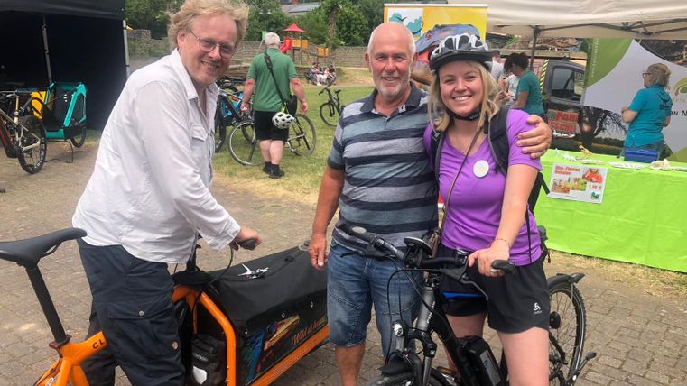 Three people are smiling in front of a bicycle and a tent. In the background, more bicycles and stalls can be seen.