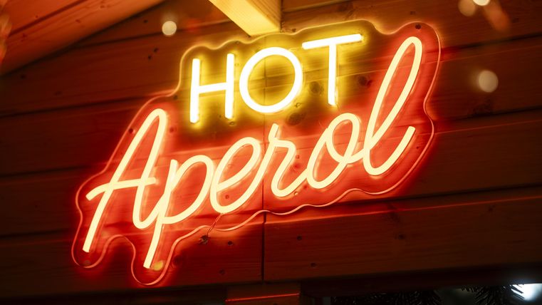 Ein leuchtendes Neonschild mit der Aufschrift "Hot Aperol" in warmen Farben. Das Schild ist in einer gemütlichen Umgebung angebracht.