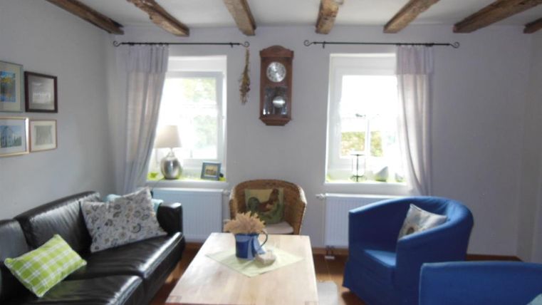Ein gemütliches Wohnzimmer mit Holzdeckenbalken. Es gibt ein schwarzes Sofa, zwei blaue Sessel und einen Holztisch in der Mitte.