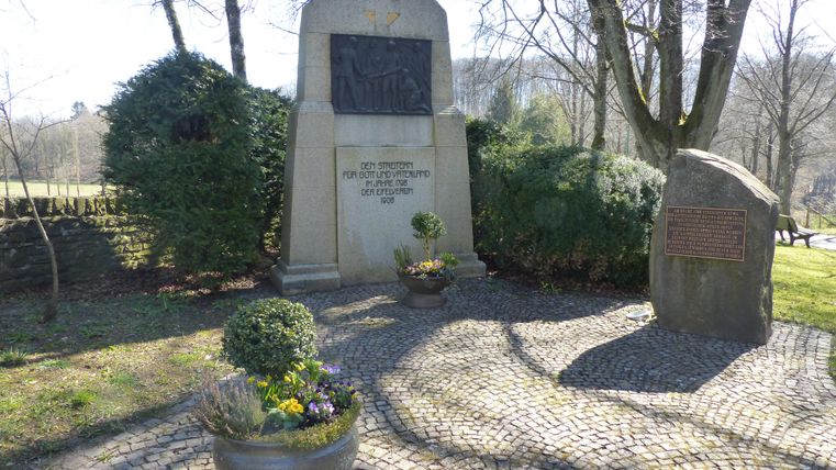 Een historisch gebouw met torens staat voor een beboste heuvelachtergrond. Het plein heeft een rustige sfeer en wordt omringd door enkele auto's.