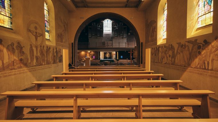 View through the nave of a church with empty rows of wooden pews. Depictions of the Stations of the Cross and colorful stained glass windows can be seen on the walls. The chancel with organ and tabernacle is visible in the background.