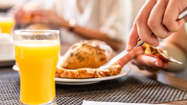 Een glas sinaasappelsap staat op de tafel naast een beboterd broodje. Een hand snijdt het broodje, terwijl op de achtergrond meer personen te zien zijn.