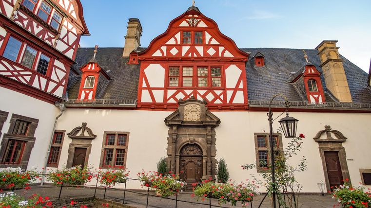 Un bâtiment historique avec une structure à colombages rouge marquante. La cour est entourée de fleurs colorées.