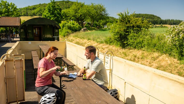 Ein Paar sitzt an einem Tisch im Freien und genießt ein Picknick. Im Hintergrund sind grüne Bäume und ein blauer Himmel zu sehen.