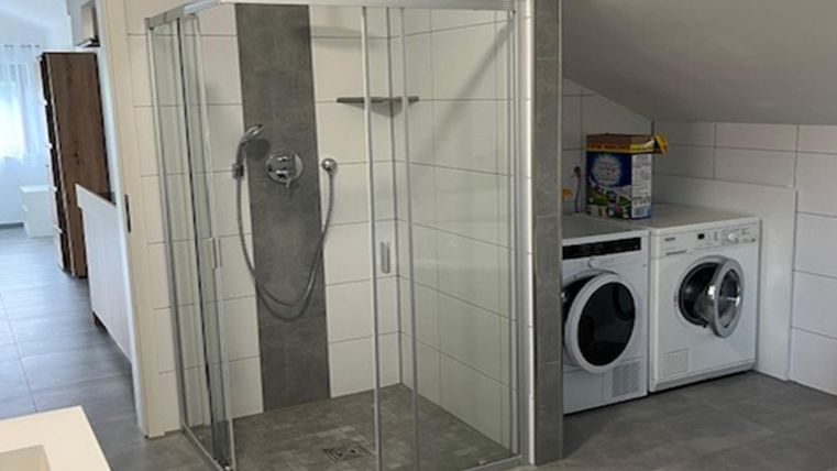 Une salle de bain moderne avec une douche à porte en verre et une machine à laver à côté d'un sèche-linge. Le sol est carrelé de carreaux gris et les murs sont carrelés en clair.
