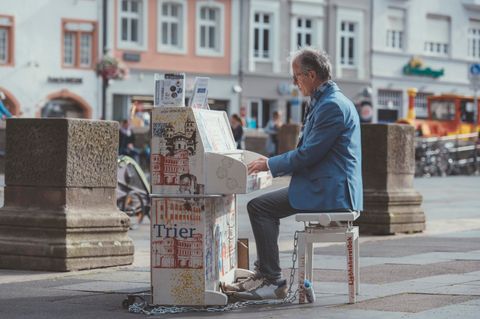 A man is playing the piano in a busy square. In the background, historical buildings and bicycles can be seen.