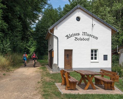 Bâtiment blanc avec l'inscription 'Petit Musée de Bolsdorf'. Deux cyclistes sur un chemin à côté. Table en bois et bancs au premier plan.