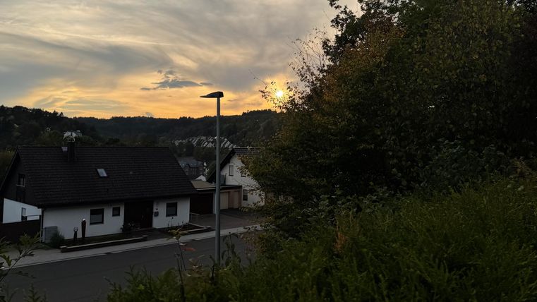 A quiet evening view with houses and trees in the foreground. The sun sets behind the hills and gently colors the sky.
