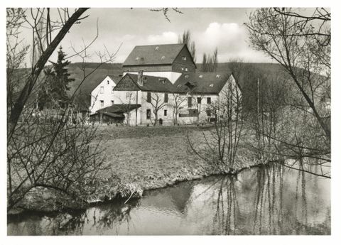Ein historisches Gebäude am Ufer eines ruhigen Flusses, umgeben von Bäumen. Die Landschaft wirkt friedlich und ländlich.