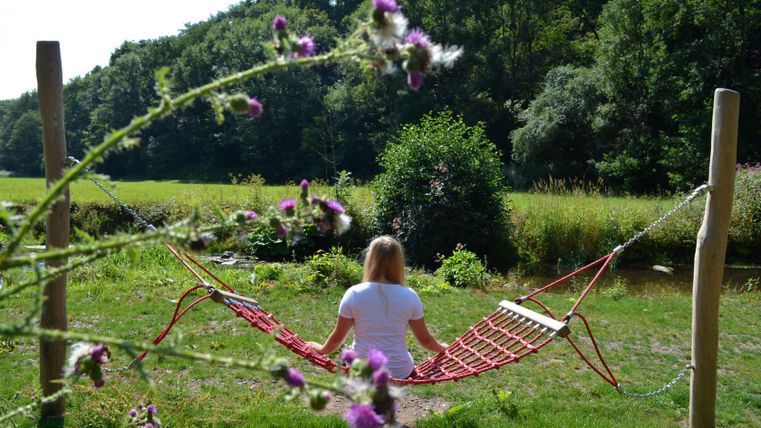 Person sits in a red hammock in a green park, surrounded by trees and flowers.