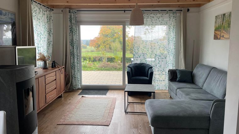 Ein gemütliches Wohnzimmer mit einem grauen Sofa und einem kleinen Tisch. Große Fenster sorgen für viel Licht und Blick auf den Garten.