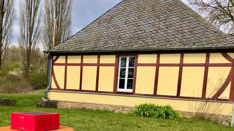 Een historisch gebouw met een schuin dak en kleurrijke muren staat op de achtergrond. Op de voorgrond zie je een rode doos op een oranje vloer.