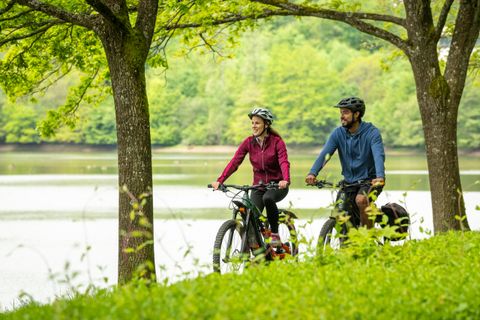 Two people ride bicycles along a lake surrounded by green nature.