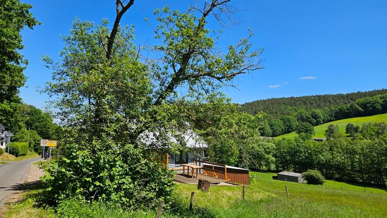 A green landscape with a large tree and a house in the background. The surroundings consist of meadows and gentle hills under a clear blue sky.
