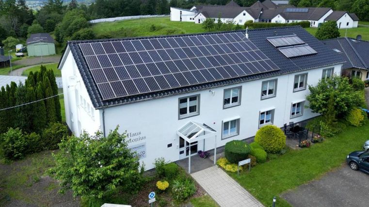 Ein modernes Gebäude mit Solarpanels auf dem Dach. Der Eingangsbereich ist von Pflanzen und Grünflächen umgeben.
