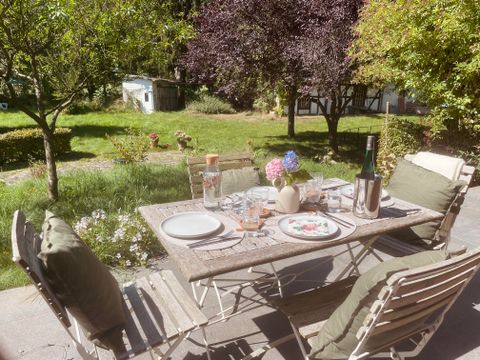 Een mooi gedekte tafel met servies en een fles wijn staat buiten. Op de achtergrond zijn bomen en een verzorgde tuin te zien.