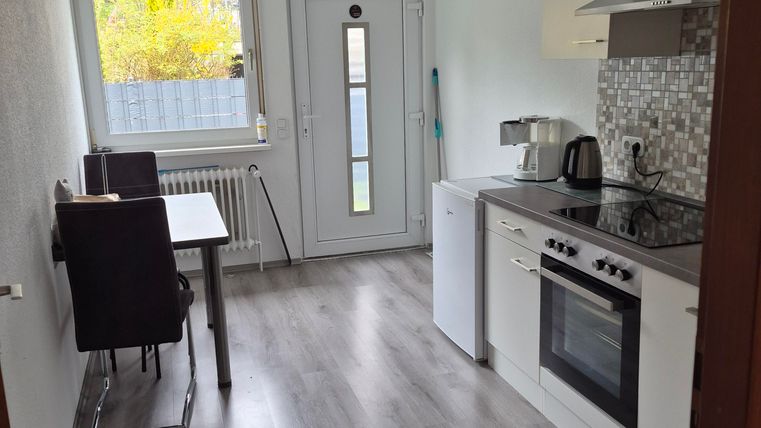 Een moderne keuken met een fornuis, een gootsteen en een koel-vriescombinatie. Er is een tafel met stoelen en een groot raam dat daglicht binnenlaat.