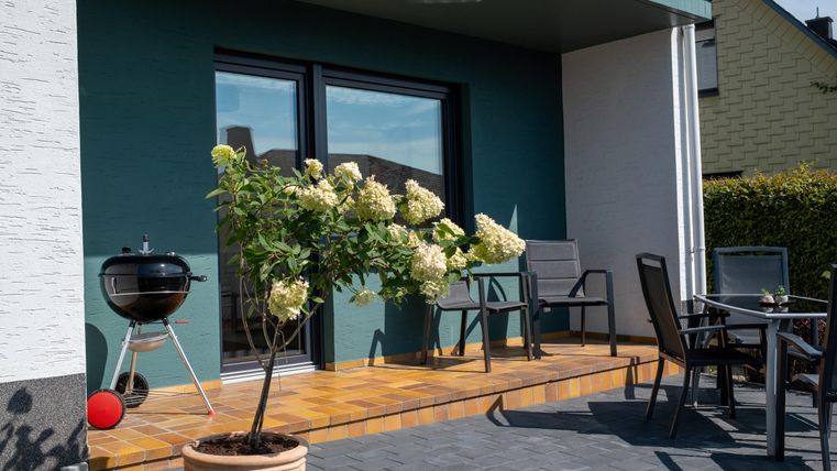 A cozy terrace with modern furniture and a grill. In front of the terrace, white potted plants are blooming.